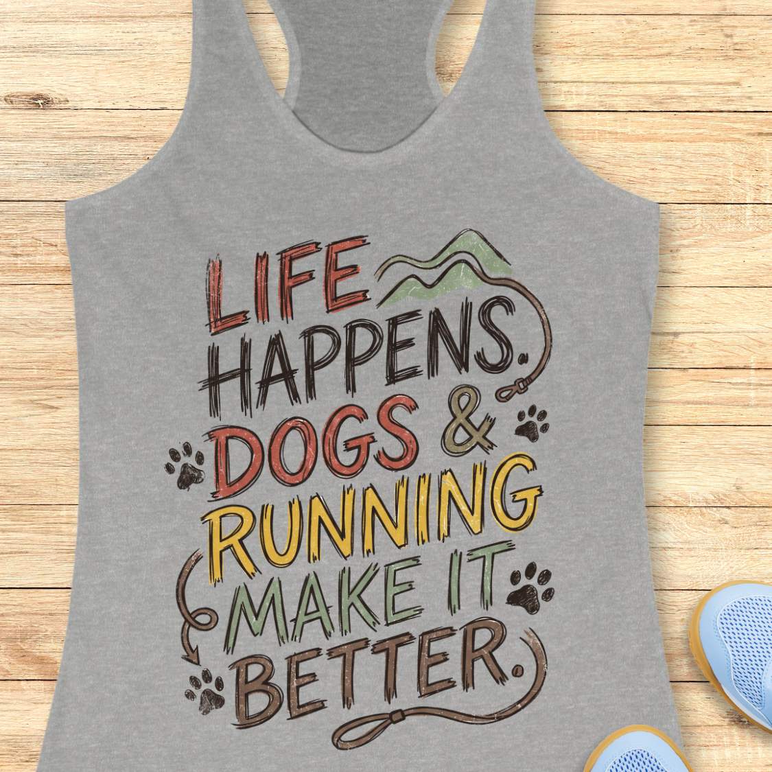 A heather gray racerback tank top laid flat on a light wood background, featuring a colorful graphic that reads 'LIFE HAPPENS. DOGS & RUNNING MAKE IT BETTER.