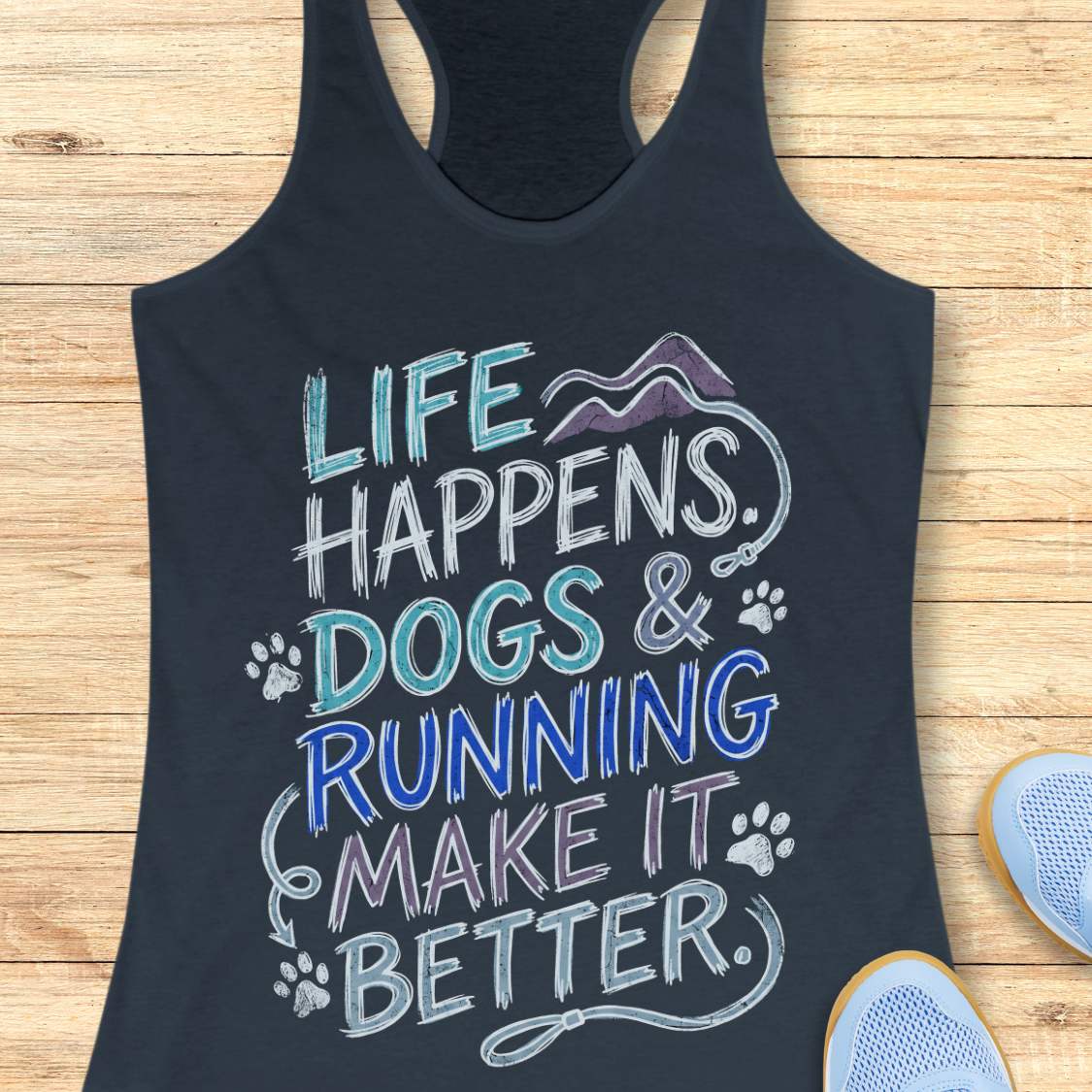 A indigo racerback tank top laid flat on a light wood surface, featuring a colorful graphic that reads 'LIFE HAPPENS. DOGS & RUNNING MAKE IT BETTER.