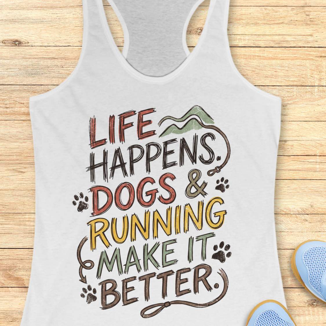 A white racerback tank top laid flat on a light wood background, featuring a colorful graphic that reads 'LIFE HAPPENS. DOGS & RUNNING MAKE IT BETTER.