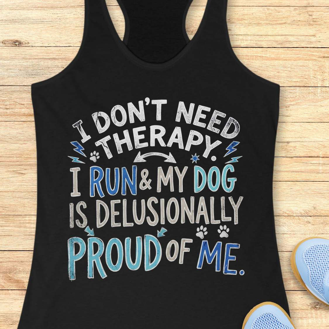A black racerback tank top laid flat on a light wood surface, featuring the text 'I DON'T NEED THERAPY. I RUN & MY DOG IS DELUSIONALLY PROUD OF ME.