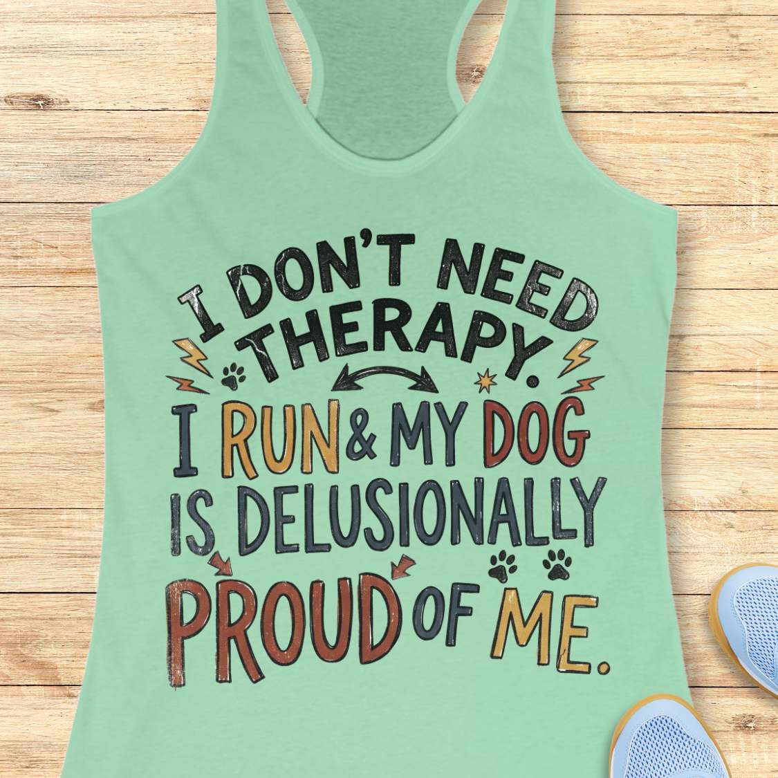 A mint green racerback tank top laid flat on a light wood background, featuring a colorful graphic text that reads 'I DON'T NEED THERAPY. I RUN & MY DOG IS DELUSIONALLY PROUD OF ME.