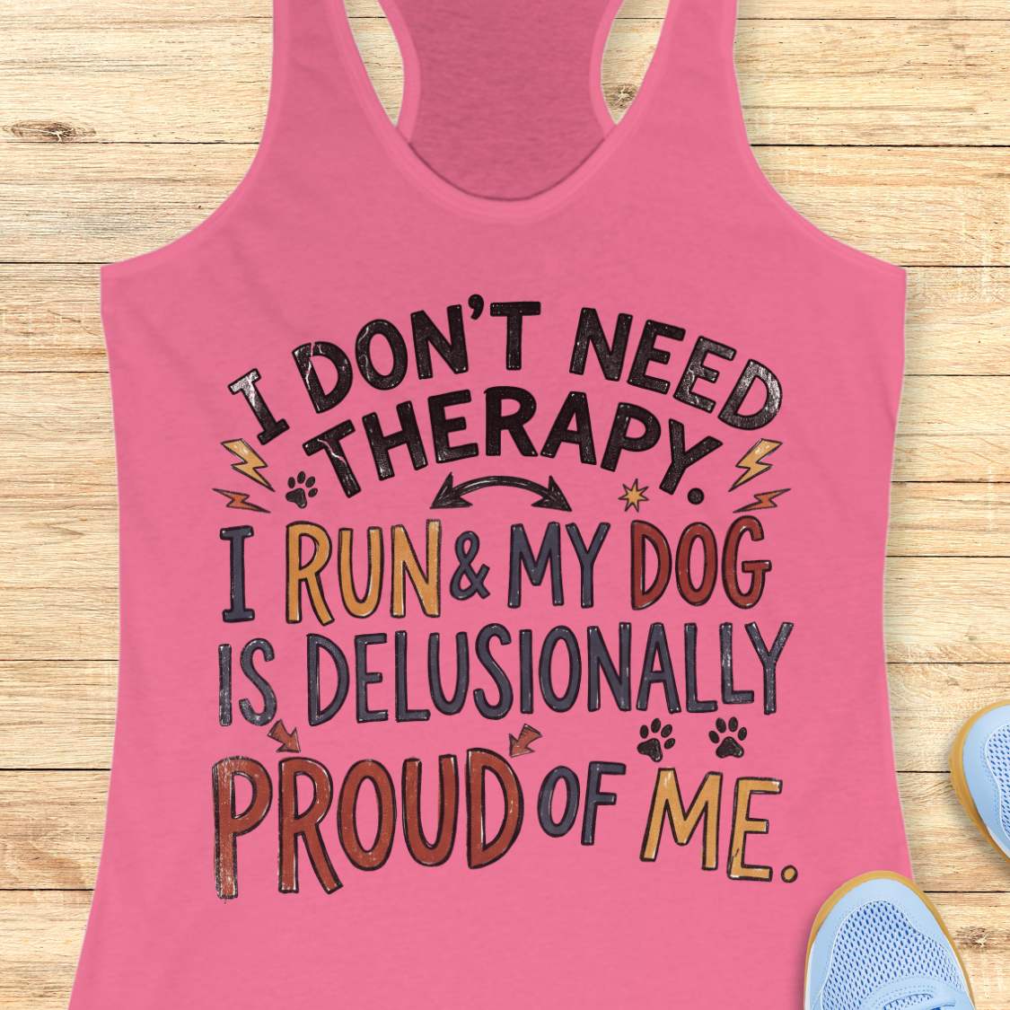 A pink racerback tank top laid flat on a light wood background, featuring a humorous graphic that reads 'I DON'T NEED THERAPY. I RUN & MY DOG IS DELUSIONALLY PROUD OF ME.