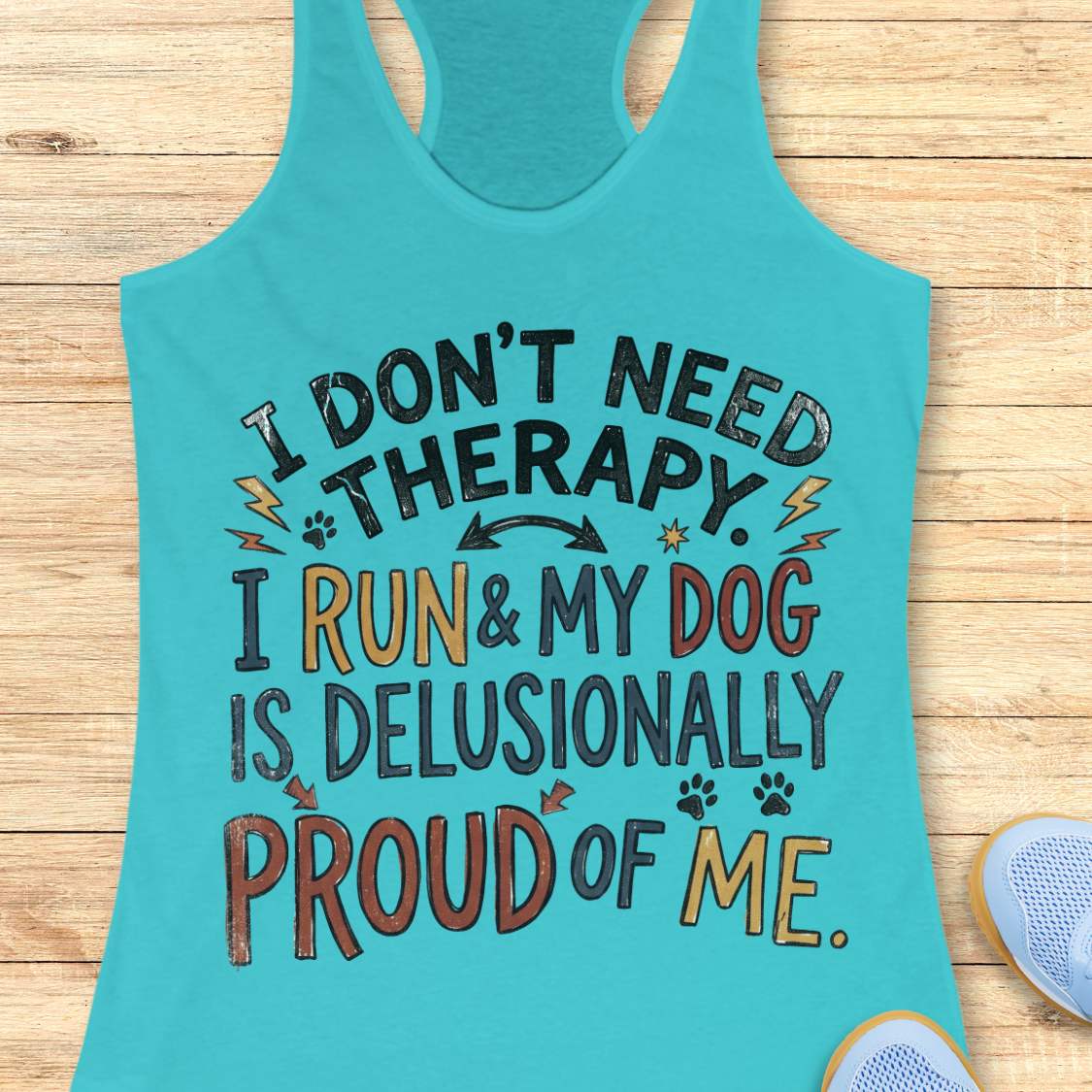 A turquoise racerback tank top laid flat on a light wood surface, featuring a colorful graphic text that reads 'I DON'T NEED THERAPY. I RUN & MY DOG IS DELUSIONALLY PROUD OF ME.