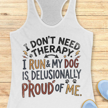 A white racerback tank top laid flat on a light wood plank background, featuring a colorful graphic text that reads 'I DON'T NEED THERAPY. I RUN & MY DOG IS DELUSIONALLY PROUD OF ME.