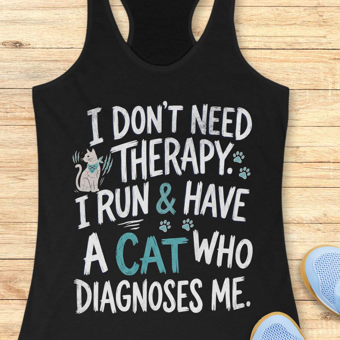 A black racerback tank top laid flat on a light wood surface, featuring a humorous graphic print with a small cat illustration and the text 'I DON'T NEED THERAPY. I RUN & HAVE A CAT WHO DIAGNOSES ME.