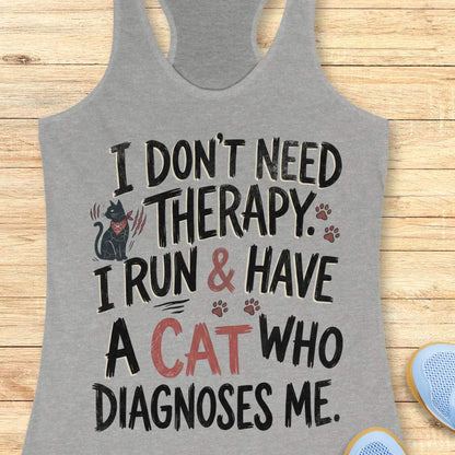 A heather gray racerback tank top laid flat on a light wood background, featuring a humorous graphic print that reads 'I DON'T NEED THERAPY. I RUN & HAVE A CAT WHO DIAGNOSES ME.