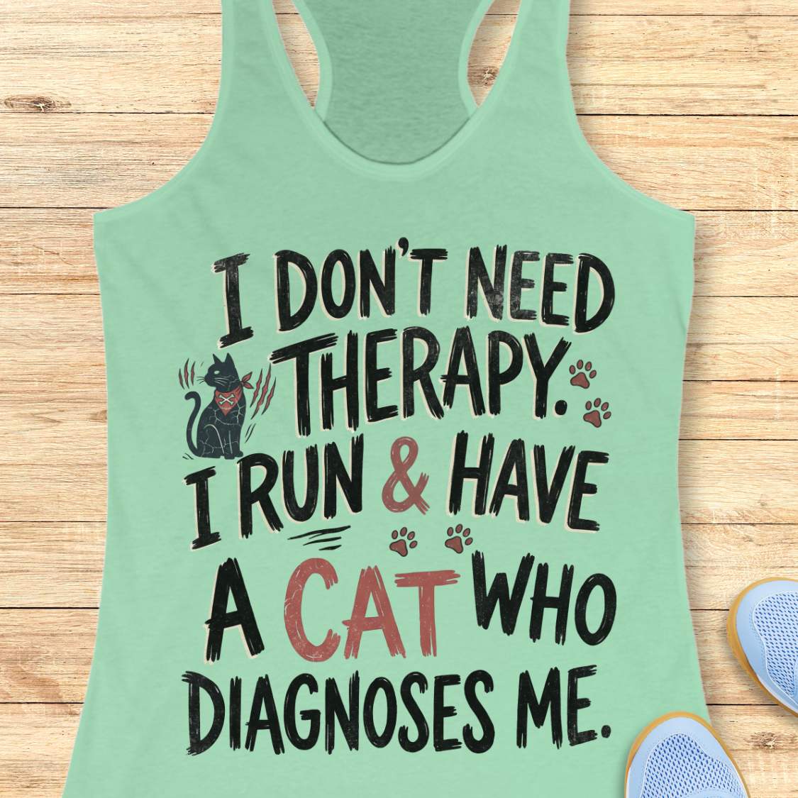 A mint green racerback tank top with a humorous graphic print, laid flat on a light wood background. The print features a black cat illustration and the text 'I DON'T NEED THERAPY.