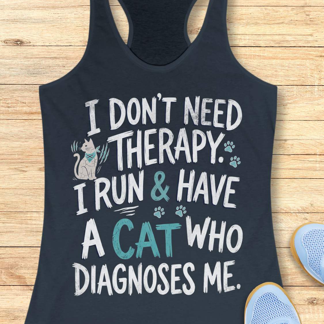 A indigo racerback tank top laid flat on a light wood background, featuring a humorous graphic that reads 'I DON'T NEED THERAPY. I RUN & HAVE A CAT WHO DIAGNOSES ME.