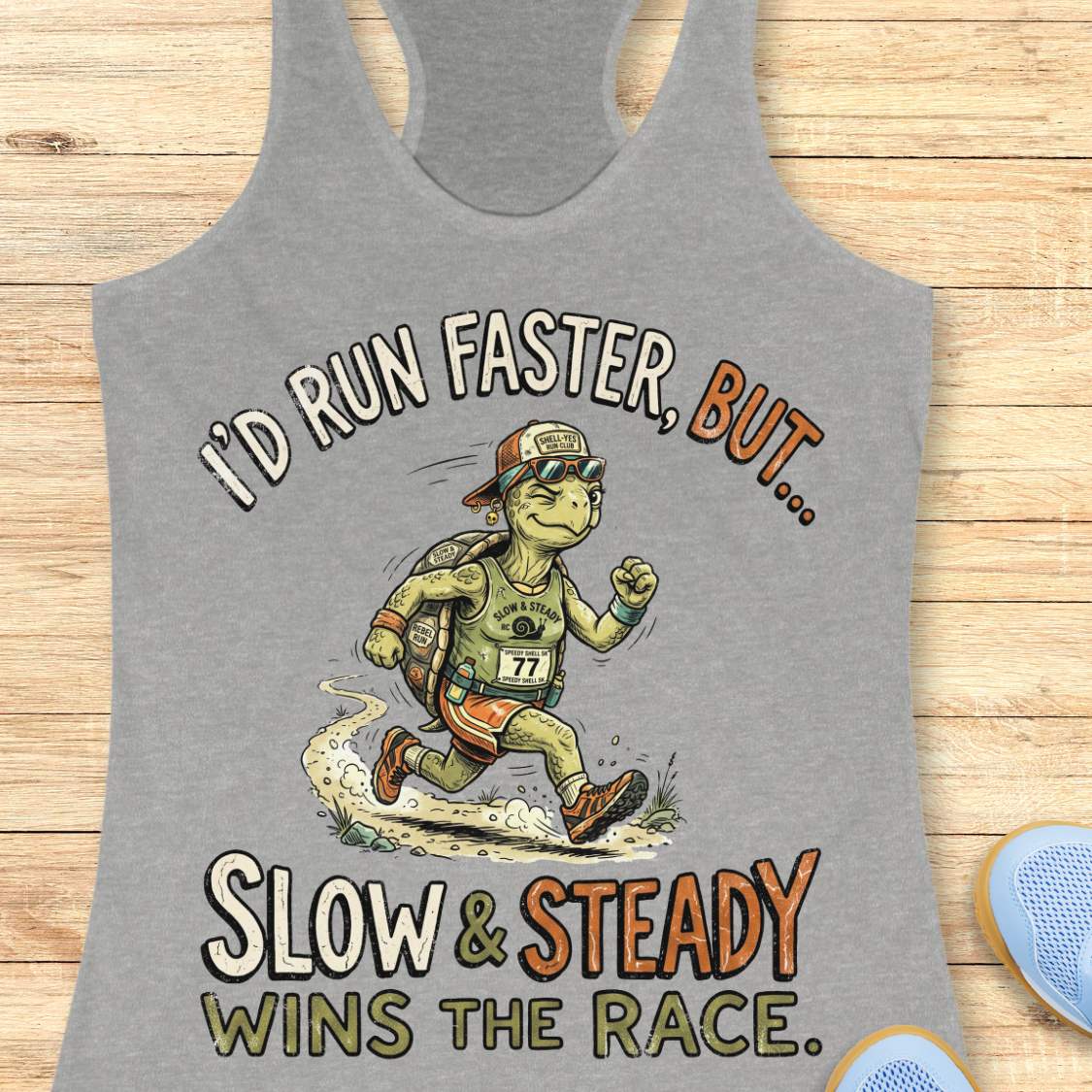 A heather gray racerback tank top laid flat on a light wood surface, featuring a large graphic of a turtle runner with the text 'I'D RUN FASTER, BUT.