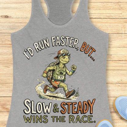 A heather gray racerback tank top laid flat on a light wood surface, featuring a large graphic of a turtle runner with the text 'I'D RUN FASTER, BUT.