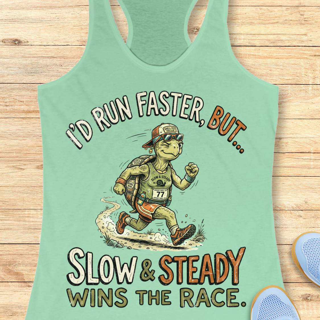 A mint green racerback tank top laid flat on a light wood background, featuring a cartoon turtle runner graphic and the text 'I'D RUN FASTER, BUT.