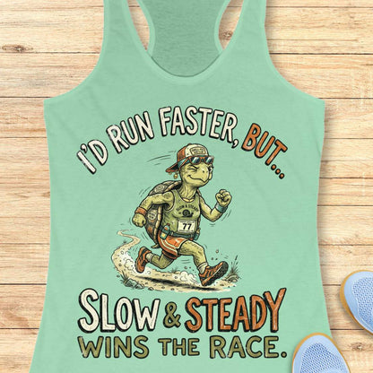 A mint green racerback tank top laid flat on a light wood background, featuring a cartoon turtle runner graphic and the text 'I'D RUN FASTER, BUT.