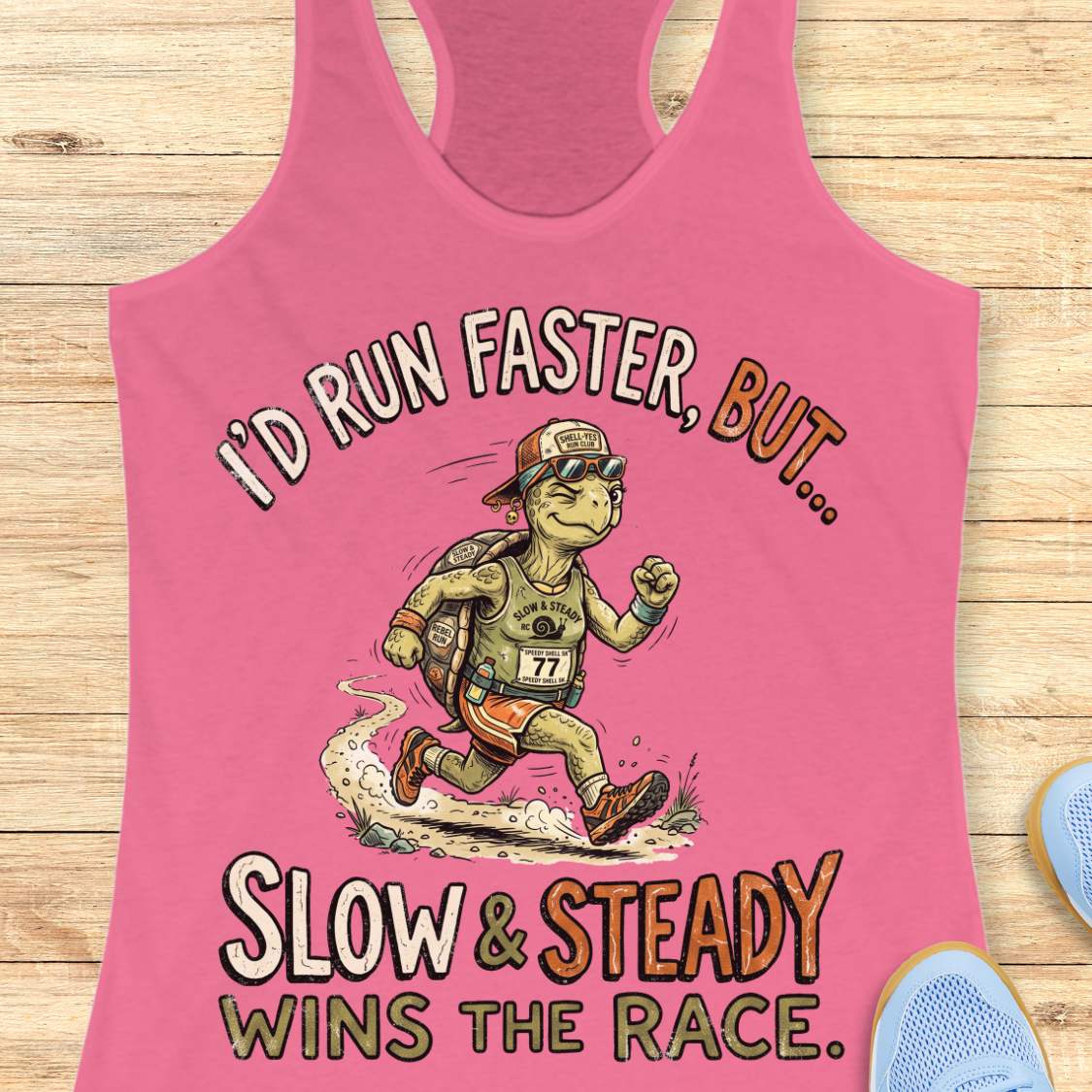 A pink racerback tank top laid flat on a light wood background, featuring a cartoon turtle runner graphic and the text 'I'D RUN FASTER, BUT.