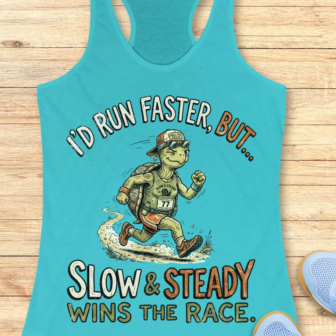 A turquoise racerback tank top laid flat on a light wood surface, featuring a cartoon turtle runner graphic with the text 'I'D RUN FASTER, BUT.