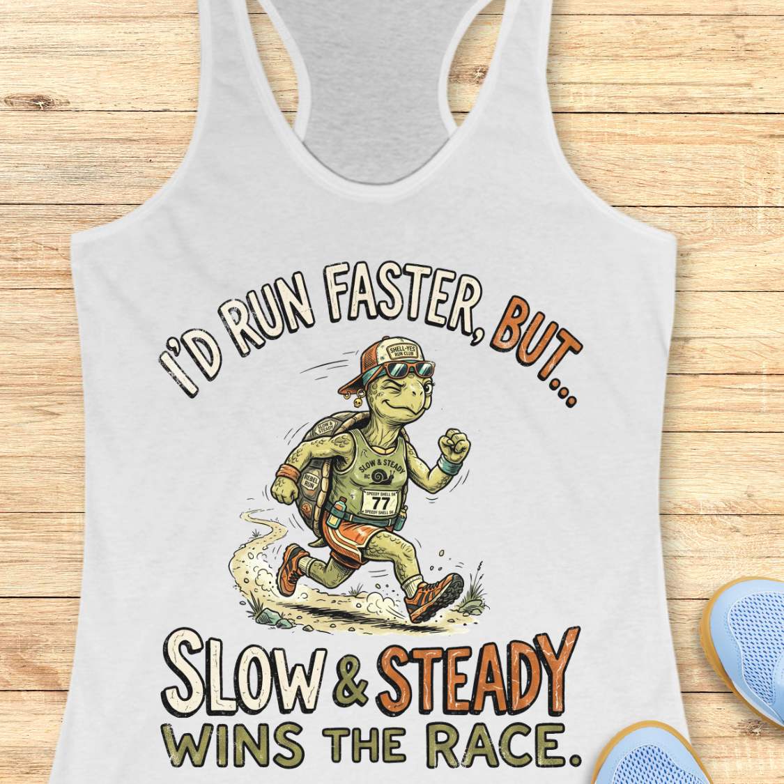 A white racerback tank top laid flat on a light wood background, featuring a cartoon turtle runner graphic with the text 'I'D RUN FASTER, BUT.