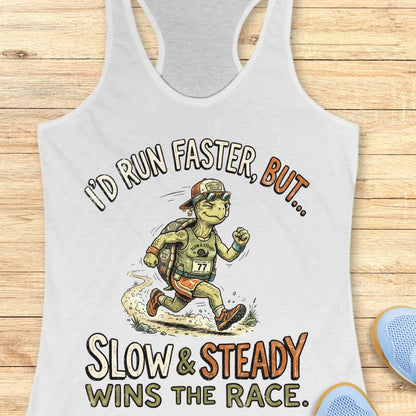 A white racerback tank top laid flat on a light wood background, featuring a cartoon turtle runner graphic with the text 'I'D RUN FASTER, BUT.
