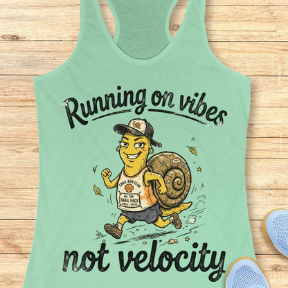 A mint green racerback tank top laid flat on a light wood background, featuring a cartoon illustration of a woman running while carrying a large snail shell, with the text 'Running on vibes not velocity' printed in black.