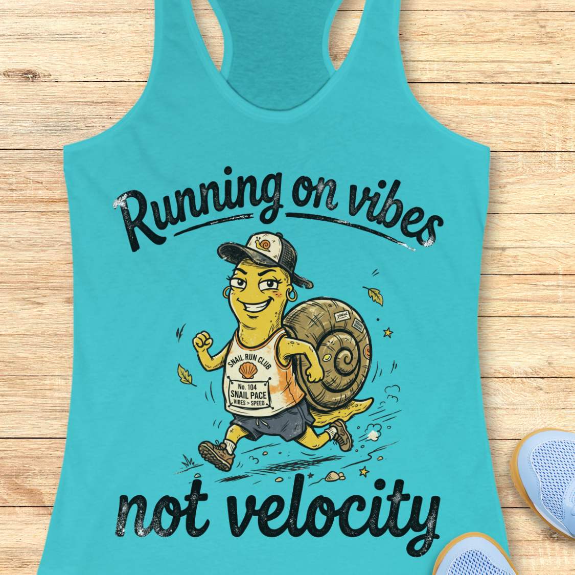 A turquoise racerback tank top laid flat on a light wood surface, featuring a cartoon runner carrying a large snail shell with the text 'Running on vibes not velocity.' A partial blue shoe appears at the lower right edge.