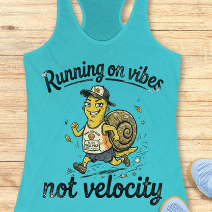 A turquoise racerback tank top laid flat on a light wood surface, featuring a cartoon runner carrying a large snail shell with the text 'Running on vibes not velocity.' A partial blue shoe appears at the lower right edge.