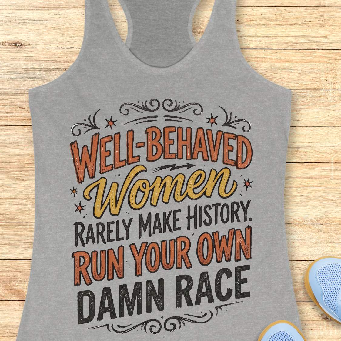 A heather gray racerback tank top with a graphic text print reading 'WELL-BEHAVED Women RARELY MAKE HISTORY. RUN YOUR OWN DAMN RACE' in orange, yellow, and black lettering, displayed on a light wood background with partial blue shoes visible at the bottom right.