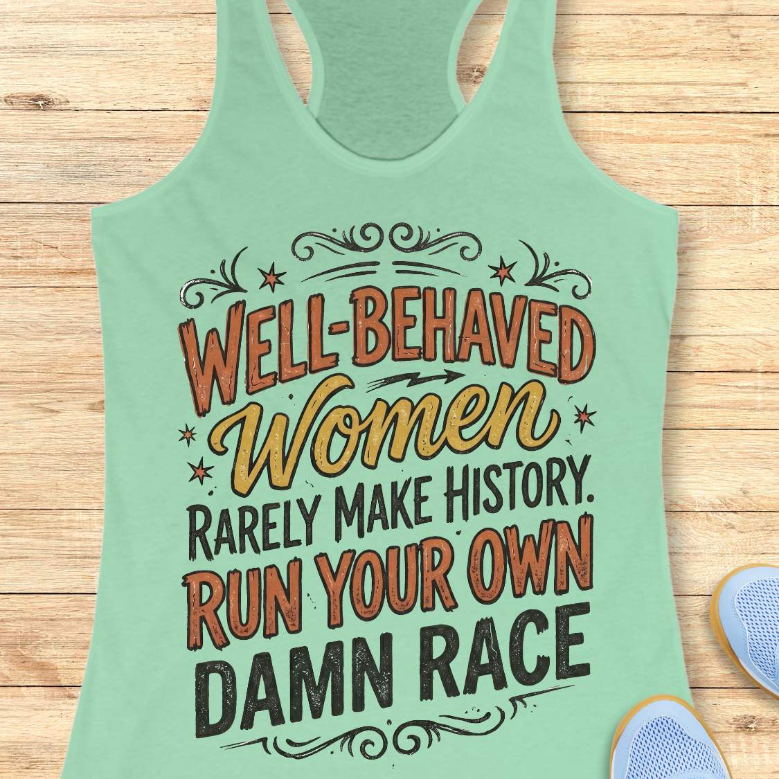 A mint green racerback tank top with a humorous graphic text reading 'WELL-BEHAVED Women RARELY MAKE HISTORY. RUN YOUR OWN DAMN RACE' in orange, yellow, and black lettering, displayed flat on a light wood background with a sneaker partially visible in the corner.
