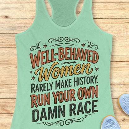 A mint green racerback tank top with a humorous graphic text reading 'WELL-BEHAVED Women RARELY MAKE HISTORY. RUN YOUR OWN DAMN RACE' in orange, yellow, and black lettering, displayed flat on a light wood background with a sneaker partially visible in the corner.