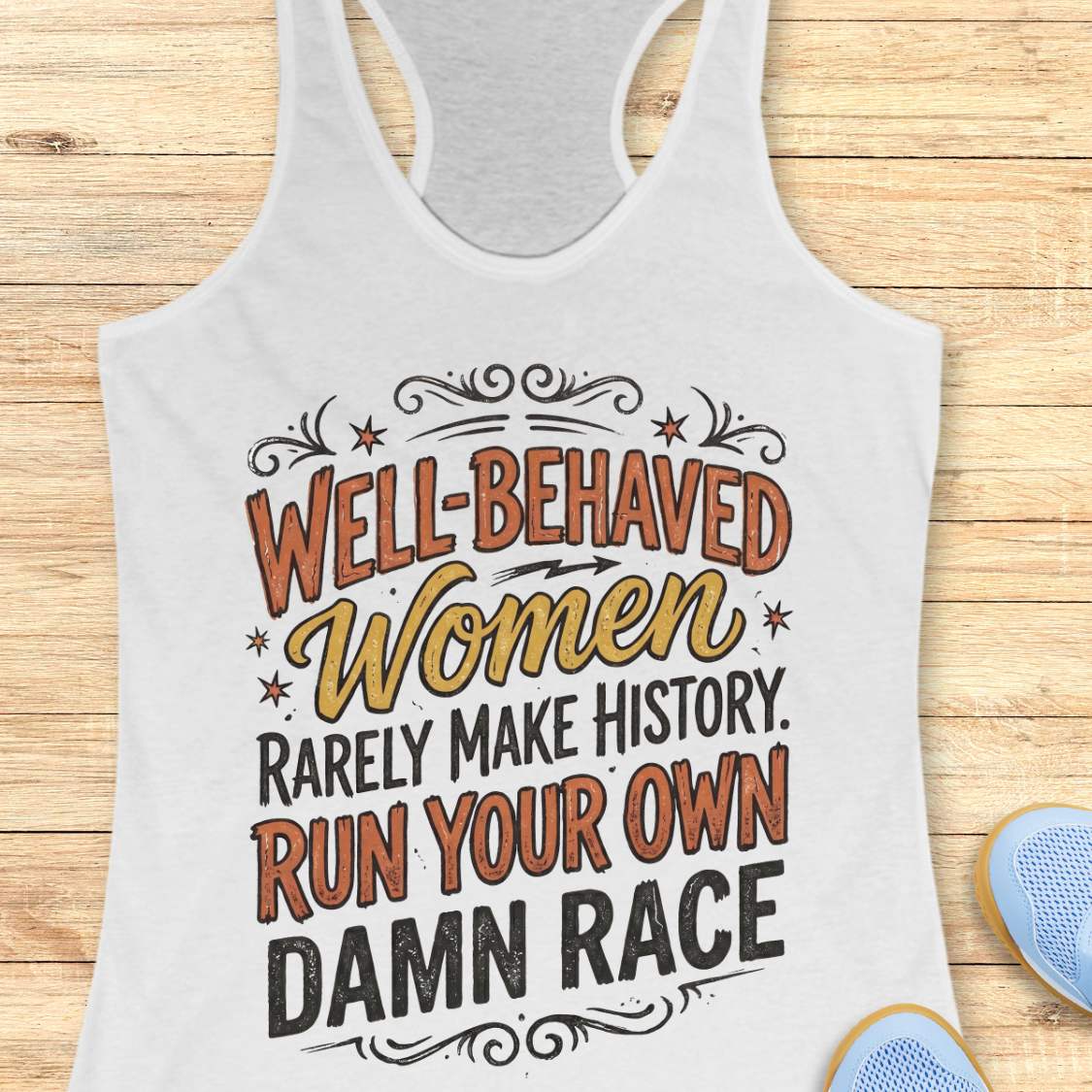 A white racerback tank top laid flat on a light wood background, featuring a motivational graphic that reads 'Well-Behaved Women Rarely Make History. Run Your Own Damn Race' in orange, yellow, and black lettering.