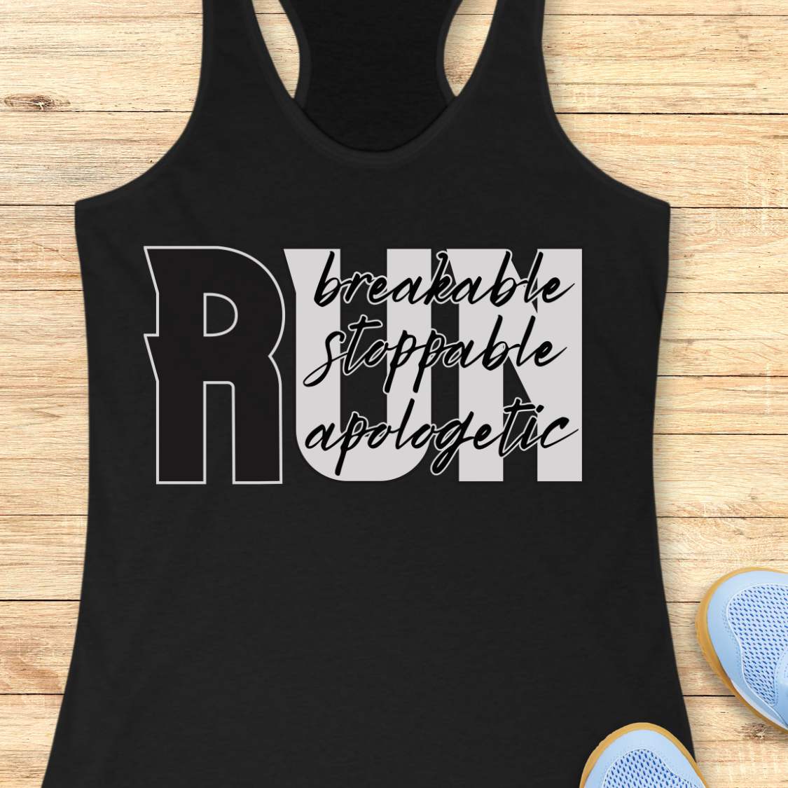 A black racerback tank top laid flat on a light wood background, featuring a large graphic that reads 'RUN' with the words 'breakable stoppable apologetic' in white and gray script across the front. Partial blue sneakers are visible at the bottom right as styling props.