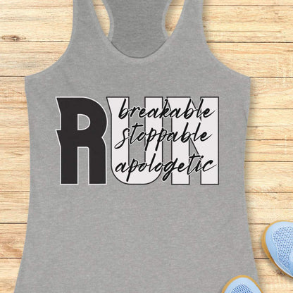 A heather gray racerback tank top laid flat on a light wood background, featuring a large graphic that reads “RUN” with the words “breakable stopable apologetic” in black script and block lettering. Partial blue shoes are visible at the bottom right as styling props.
