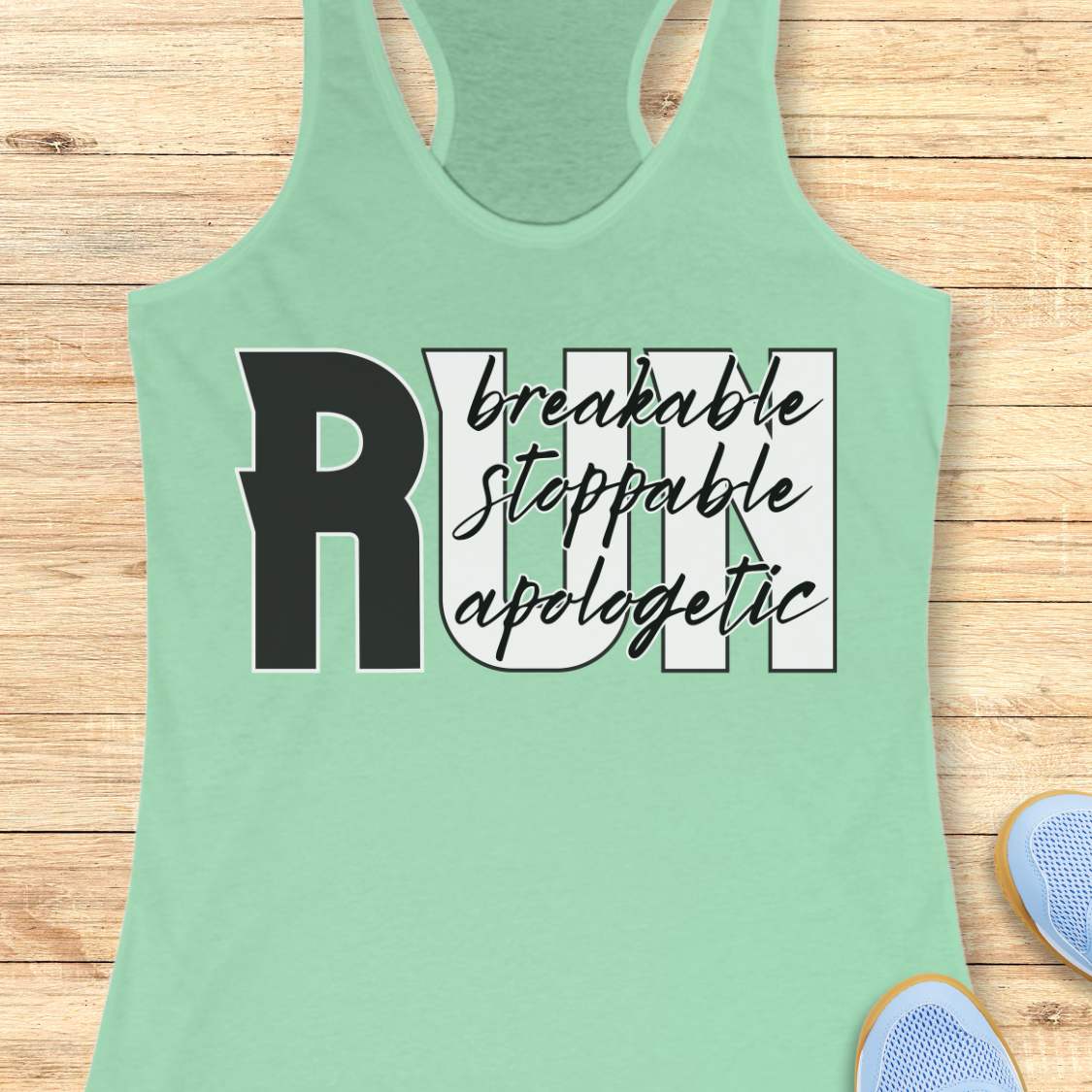 A mint green racerback tank top laid flat on a light wood background, featuring a graphic print that reads 'RUN' with 'breakable stoppable apologetic' in black script and block lettering. Partial blue sneakers are visible at the bottom right corner.