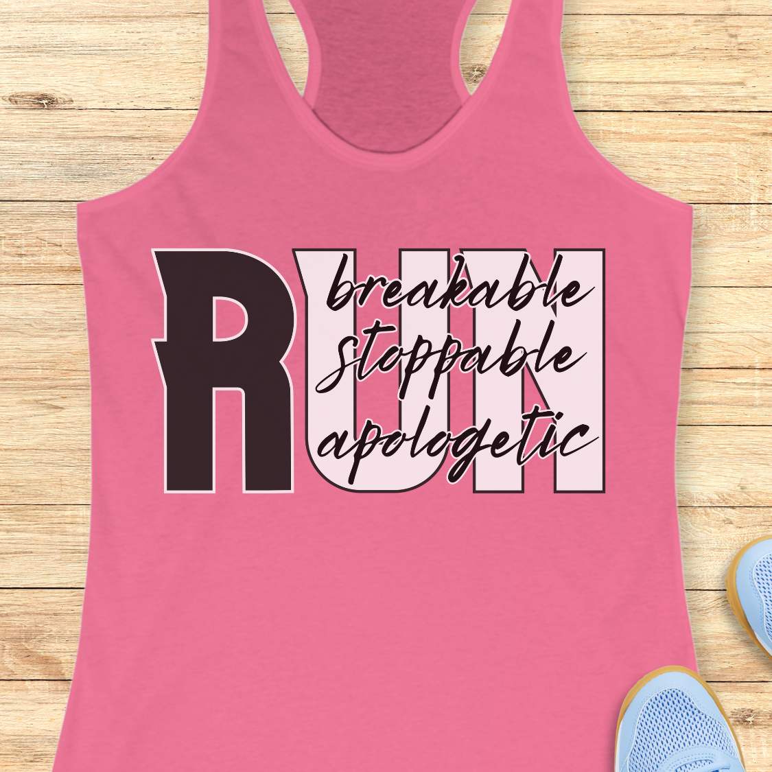 A pink racerback tank top laid flat on a light wood background, featuring a large graphic that reads “RUN” with the words “breakable stopable apologetic” in script across the letters. Partial blue shoes are visible at the bottom right as styling props.