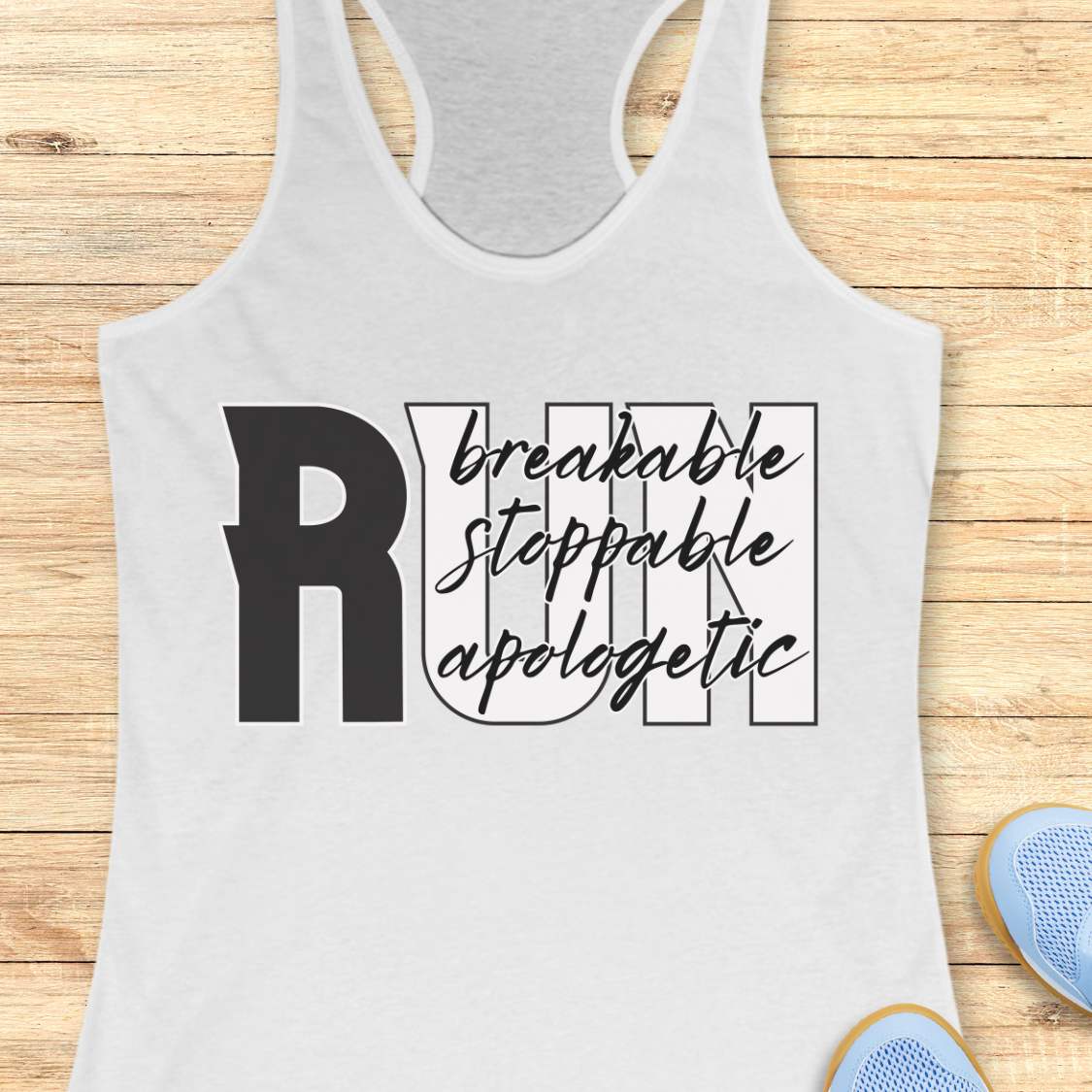 A white racerback tank top with a black graphic text print reading 'RUN breakable stoppable apologetic' laid flat on a light wood plank background. Part of a blue shoe is visible in the bottom right corner.