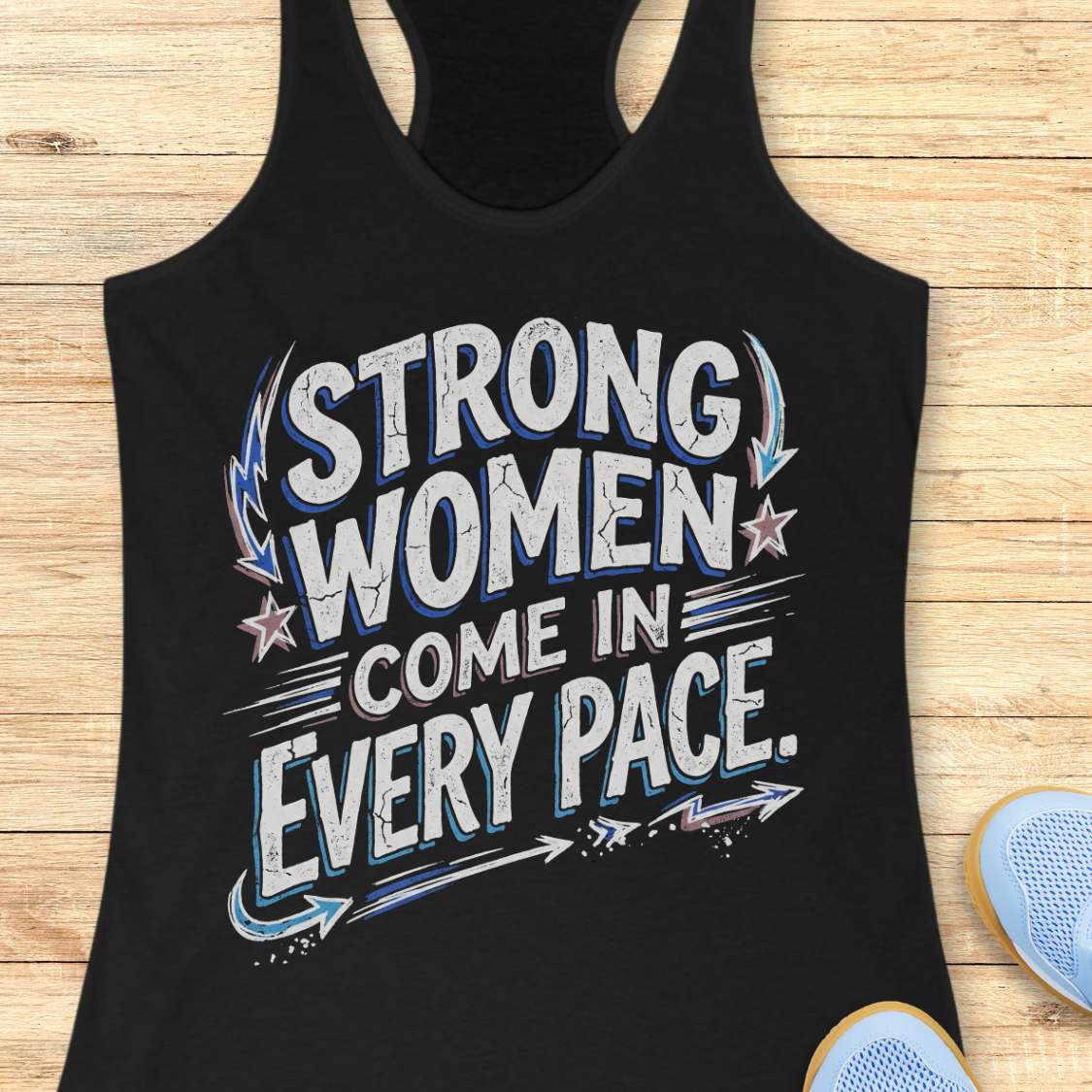 A black racerback tank top with a distressed graphic that reads 'STRONG WOMEN COME IN EVERY PACE.' in white, blue, and gray, laid flat on a light wood background with partial blue sneakers visible at the bottom right.