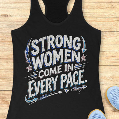 A black racerback tank top with a distressed graphic that reads 'STRONG WOMEN COME IN EVERY PACE.' in white, blue, and gray, laid flat on a light wood background with partial blue sneakers visible at the bottom right.
