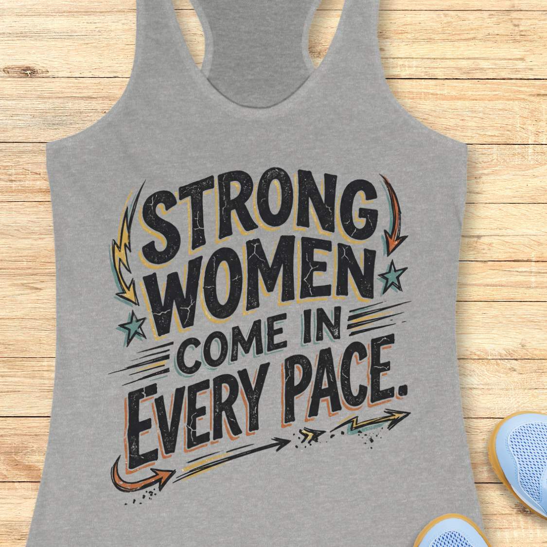 A heather gray racerback tank top laid flat on a light wood surface, featuring the distressed text 'STRONG WOMEN COME IN EVERY PACE' in black with orange and teal graphic accents. Partial blue shoes are visible at the bottom right as styling props.