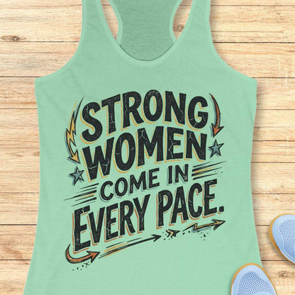 A mint green racerback tank top with a distressed black graphic that reads 'STRONG WOMEN COME IN EVERY PACE.' in stylized lettering, shown laid flat on a light wood plank background with partial sneakers visible at the edges.
