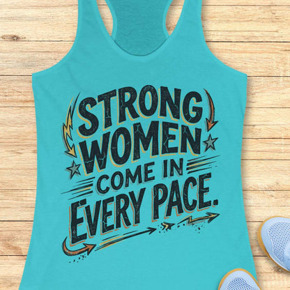 A turquoise racerback tank top laid flat on a light wood surface, featuring the printed slogan 'STRONG WOMEN COME IN EVERY PACE' in distressed black lettering with gold and dark gray arrow and star accents. Part of a white sneaker is visible at the bottom right.