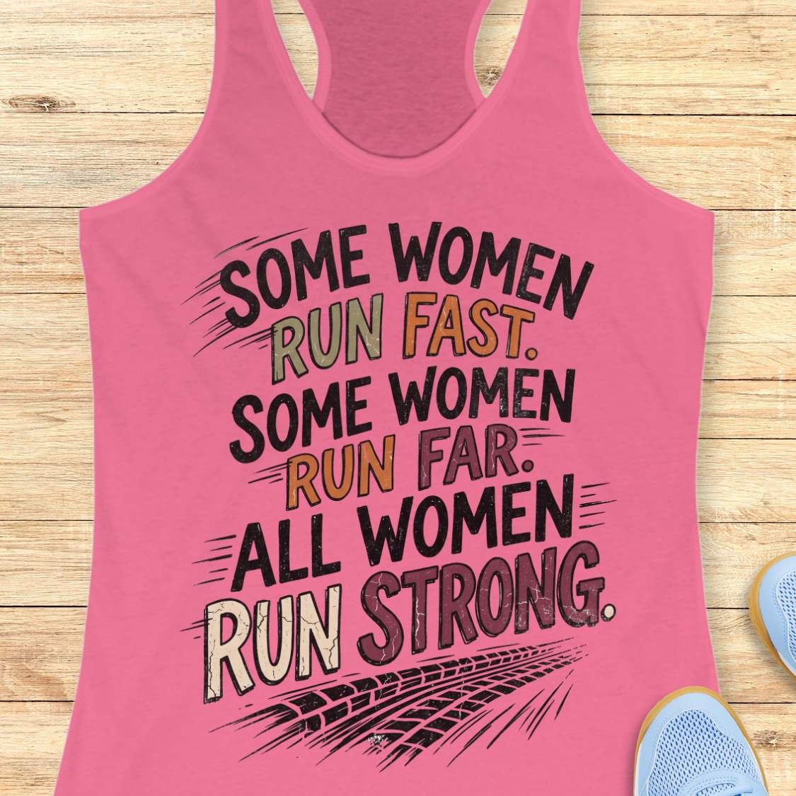 A pink racerback tank top laid flat on a light wood surface, featuring a motivational graphic with illustrated sneakers.