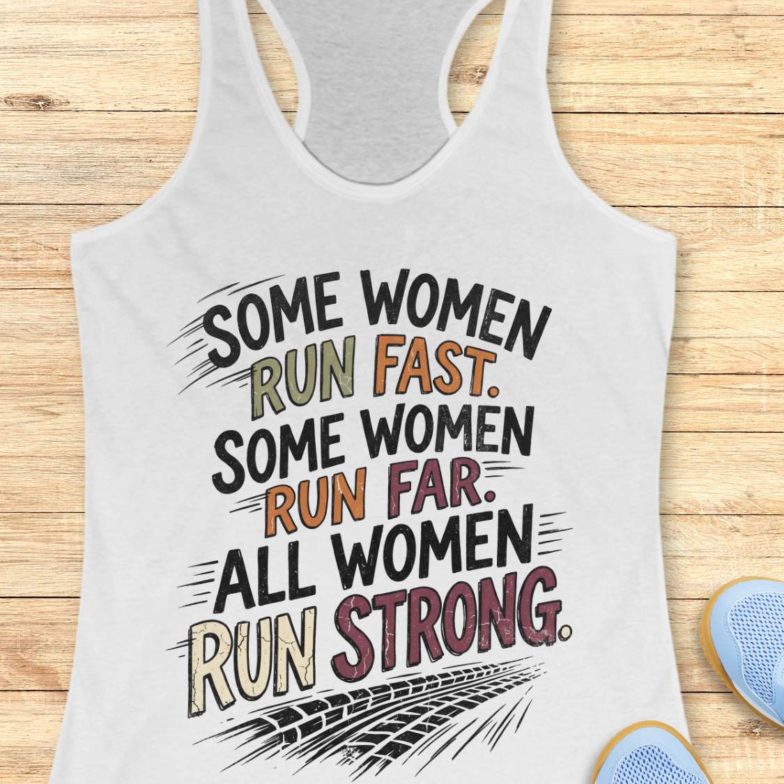 A white racerback tank top laid flat on a light wood surface, featuring a motivational graphic with illustrated sneakers.