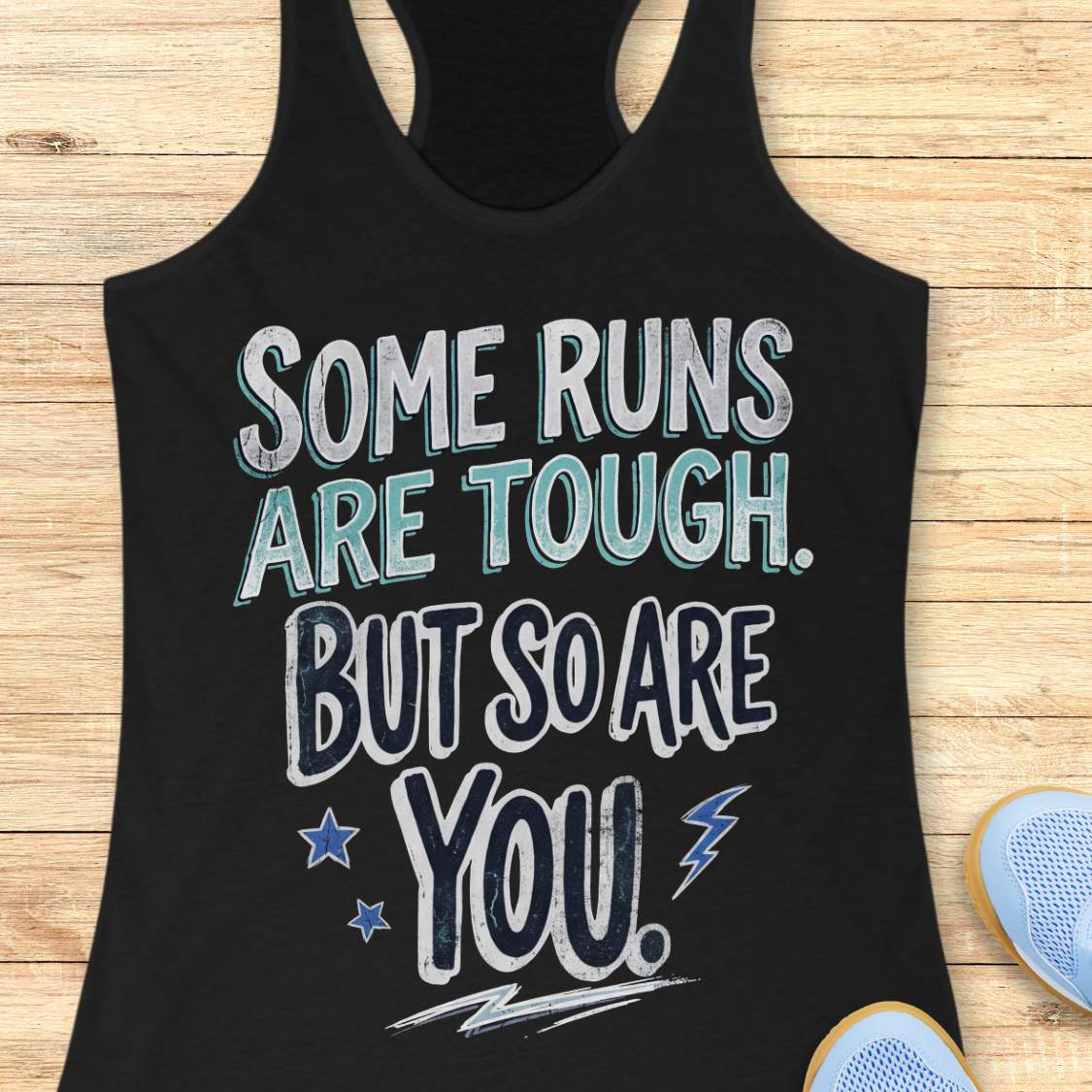 A black racerback tank top laid flat on a light wood surface, featuring a motivational graphic with illustrated sneakers.