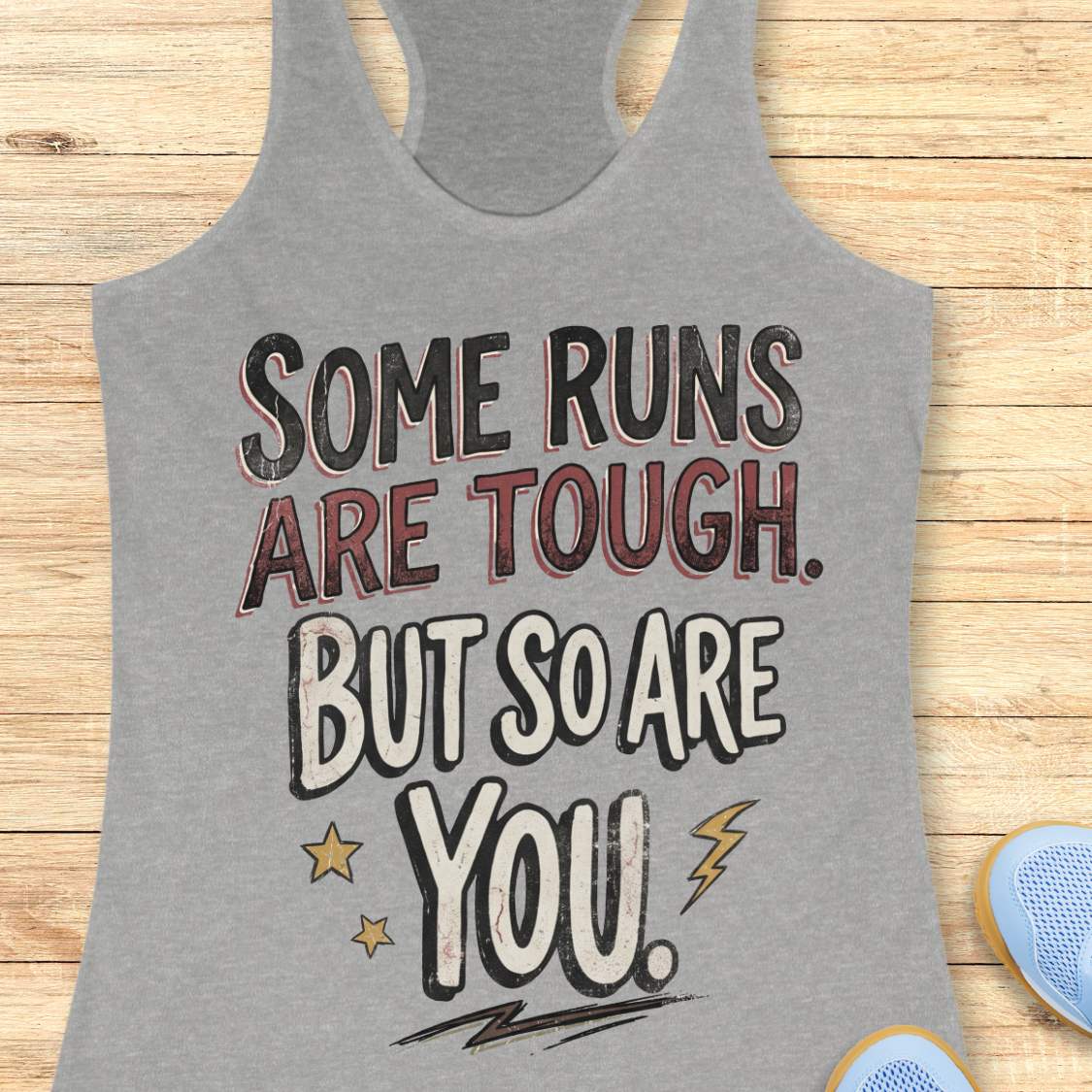 A heather grey racerback tank top laid flat on a light wood surface, featuring a motivational graphic with illustrated sneakers.