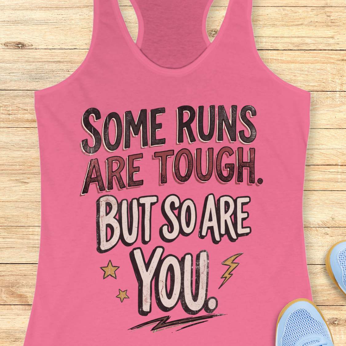 A pink racerback tank top laid flat on a light wood surface, featuring a motivational graphic with illustrated sneakers.