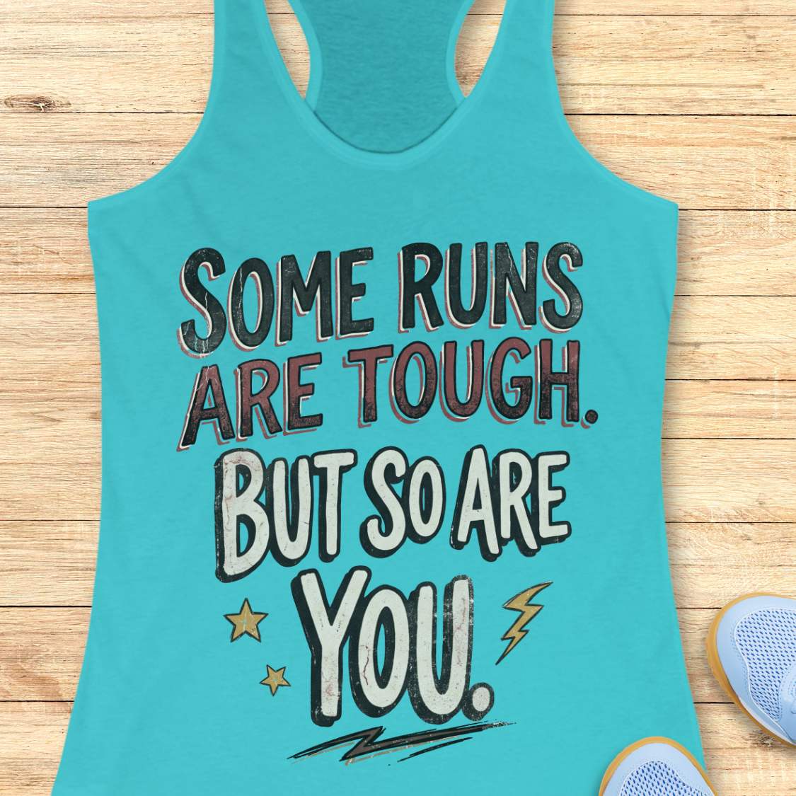 A blue racerback tank top laid flat on a light wood surface, featuring a motivational graphic with illustrated sneakers.