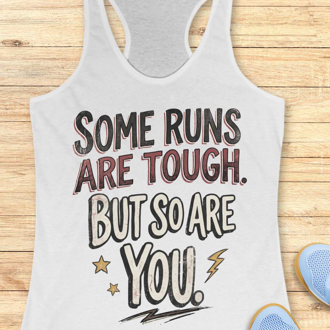 A white racerback tank top laid flat on a light wood surface, featuring a motivational graphic with illustrated sneakers.