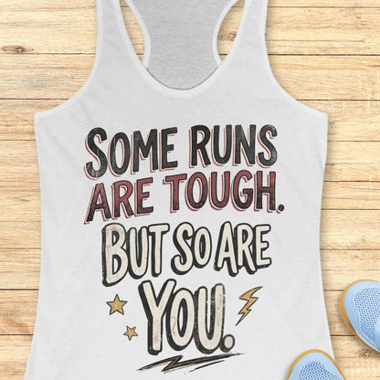 A white racerback tank top laid flat on a light wood surface, featuring a motivational graphic with illustrated sneakers.
