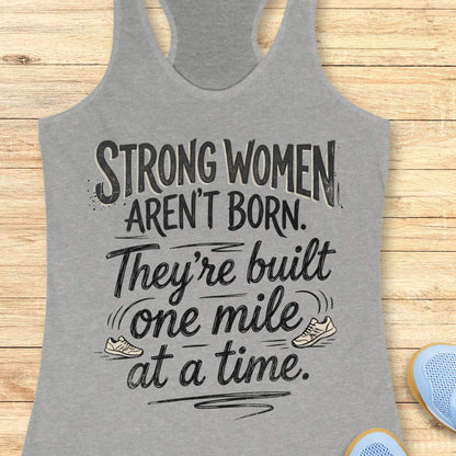 A grey racerback tank top laid flat on a light wood surface, featuring a motivational graphic with illustrated sneakers.
