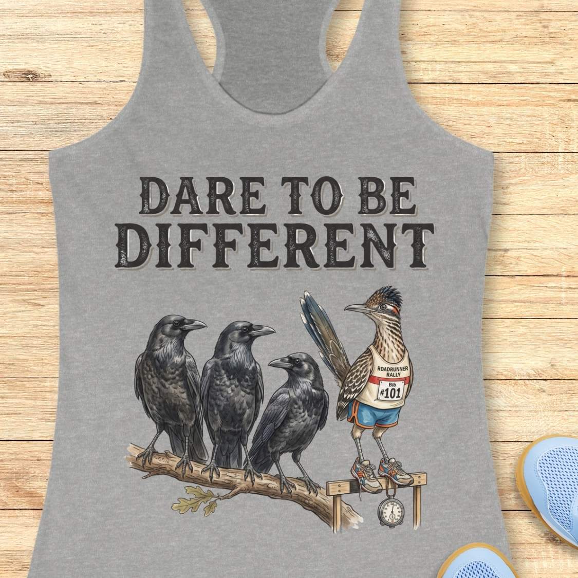 A grey racerback tank top laid flat on a light wood background, featuring a graphic print of three black crows and a roadrunner wearing a race bib with the phrase 'DARE TO BE DIFFERENT'.