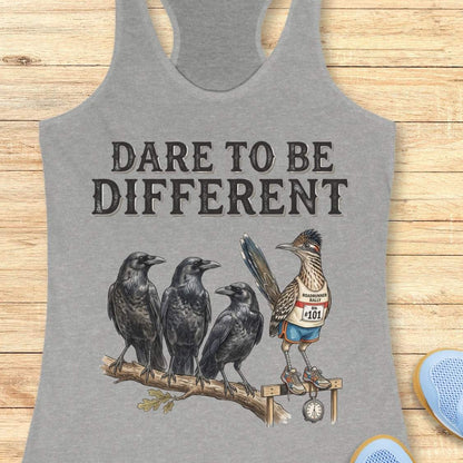 A grey racerback tank top laid flat on a light wood background, featuring a graphic print of three black crows and a roadrunner wearing a race bib with the phrase 'DARE TO BE DIFFERENT'.
