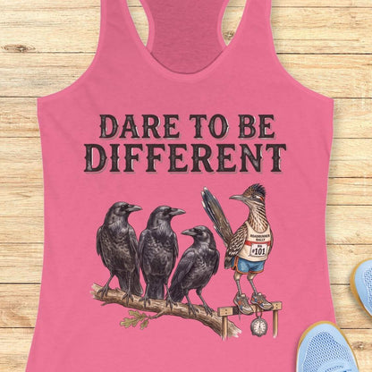 A pink racerback tank top laid flat on a light wood background, featuring a graphic print with the text 'DARE TO BE DIFFERENT' above three black crows and a roadrunner wearing a race bib.