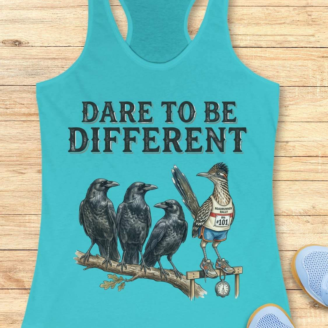 A turquoise racerback tank top laid flat on a light wood surface, featuring the printed phrase 'DARE TO BE DIFFERENT' above an illustration of three black crows and a roadrunner wearing a race bib and medal.